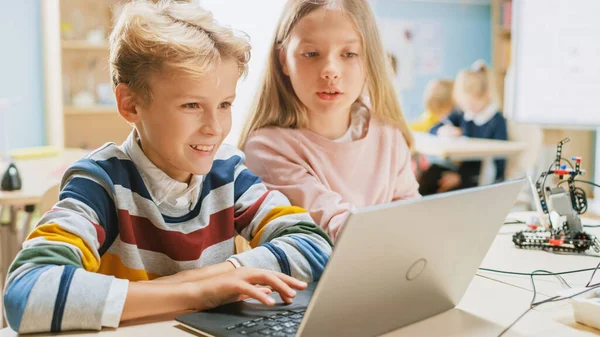 Brillante colegiala y colegial hablan y utilizan el ordenador portátil para programar software para la clase de ingeniería de robótica. Aula de Ciencias de la Escuela Primaria con Niños que Trabajan en Tecnología. Educación STEM —  Fotos de Stock