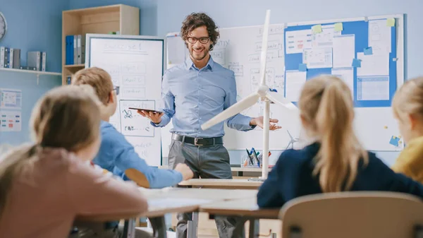 Profesor de Física de la Escuela Primaria utiliza la tableta digital interactiva para mostrar a un aula de niños inteligentes cómo funcionan las energías renovables y las turbinas eólicas. Clase de ciencias con niños escuchando atentamente —  Fotos de Stock