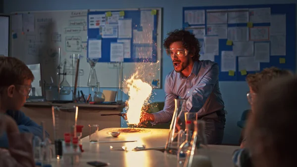 Школьный класс химии: Дети смотрят, как энтузиастичный учитель показывает научный эксперимент, подбрасывая порошок в огонь, создавая красивые фейерверки. Современное образование для детей — стоковое фото
