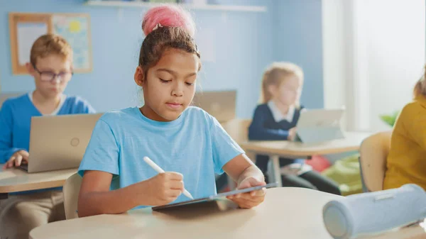Клас початкової школи комп'ютерних наук: мила дівчина використовує цифровий планшетний комп'ютер, її однокласники також працюють з ноутбуками. Діти отримують сучасну освіту в STEM, грають і навчаються — стокове фото