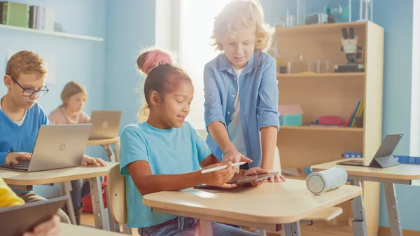 Клас початкової школи комп'ютерних наук: Смарт-дівчина використовує цифровий планшетний комп'ютер, її однокласник допомагає з завданням. Діти отримують сучасну освіту — стокове фото