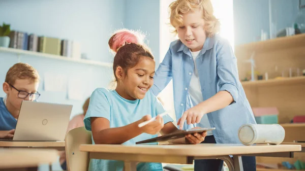 Elementary School Computer Science Class: Smart Girl utiliza Tablet Computer digital, su compañera de clase la ayuda con la tarea. Niños obteniendo educación moderna — Foto de Stock