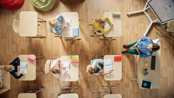 Bright Elementary School Classroom: Children Sitting at their School Desk Working, Doing Assignment, Enthusiastic Teacher Stands a the Head of the Class Explaining a Lesson. Top View Shot.