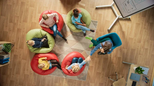 Elementary School Creativity Classroom: Kinder, die auf den Bohnensäcken sitzen, während der Lehrer ihnen den Unterricht erklärt und digitale Tablet-Computer verwendet, spielen, indem sie High Five bestehen. Schuss von oben — Stockfoto