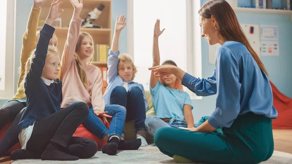 Aula de Criatividade da Escola Primária: Crianças sentadas no tapete enquanto o professor explica a lição e faz perguntas, cada criança levanta a mão com resposta. Aprendizagem em Ambiente Moderno . — Fotografia de Stock
