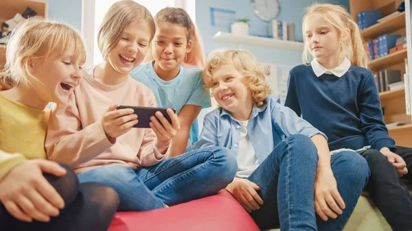 Diversi gruppi di bambini piccoli e carini seduti insieme sulle borse dei fagioli usano smartphone e parlano, divertiti. Bambini che navigano su Internet e giocano ai videogiochi online sul telefono cellulare — Foto Stock