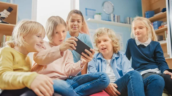 Groupe diversifié de petits enfants mignons assis ensemble sur les sacs de haricots Utilisez Smartphone et parler, amusez-vous. Enfants naviguant sur Internet et jouant à des jeux vidéo en ligne sur téléphone mobile — Photo