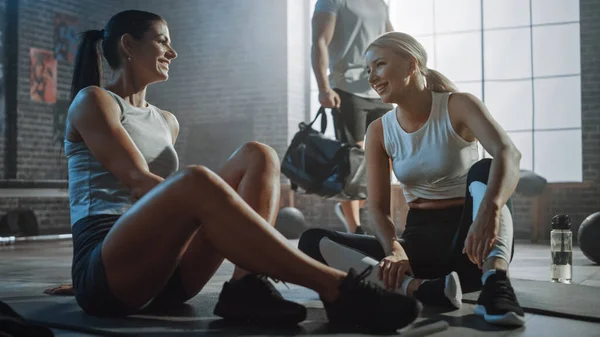 Deux belles Fit Athletic Girls Asseyez-vous sur un plancher de salle de gym industrielle Loft. Ils sont heureux avec leur programme de formation et donnent avec succès un High Five. Homme masculin fort marche en arrière-plan. — Photo