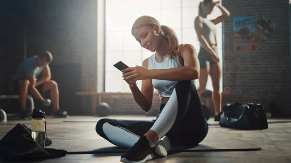Mujer joven atlética feliz y sonriente está utilizando un teléfono inteligente mientras está sentado en un piso en un gimnasio Loft. Escribe un mensaje y sonríe. Un hombre ejerce en el fondo . —  Fotos de Stock