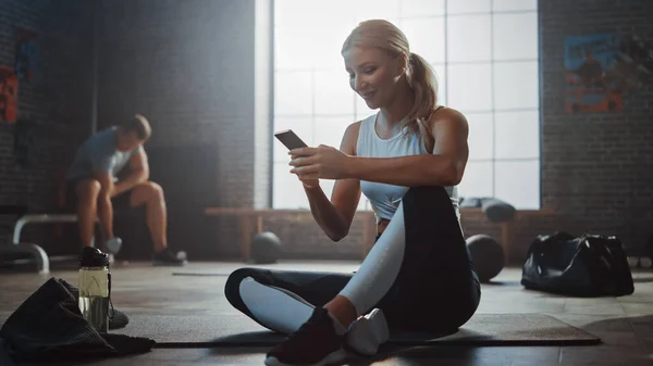 Happy and Smiling Beautiful Athletic Young Woman utilise un téléphone intelligent assis sur un plancher dans un gymnase Loft. Elle tape un message et sourit. Un homme exerce en arrière-plan. — Photo