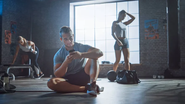 Jeune homme athlétique beau et masculin utilise un smartphone assis sur un plancher dans un gymnase Loft. Il tape un message et réfléchit. Il a de la sueur sur le front. Exercice des femmes en arrière-plan. — Photo