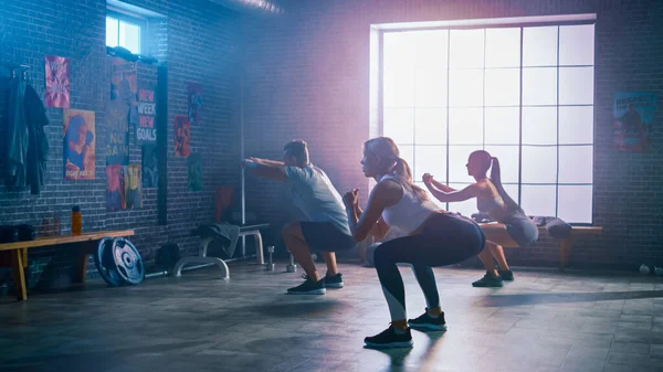 Bărbatul puternic masculin și două femei atletice se potrivesc fac exerciții squat. Se antrenează într-o sală de gimnastică cu postere motivaționale pe pereți. Cameră cu Magenta Light. Parte din echipa lor de fitness — Fotografie, imagine de stoc