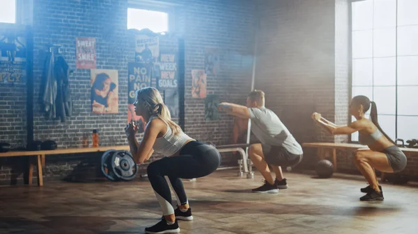 Homme Masculin Fort et Deux Fit Atletic Women font des exercices de squat. Ils s'entraînent dans un gymnase Loft avec des affiches motivationnelles sur les murs. Son Ensoleillé et Chambre a Lumière Chaude. Faire partie de leur équipe Fitness. — Photo