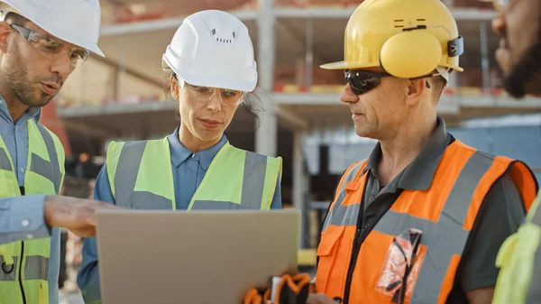 Equipo Diverso de Especialistas Utilice Computadora Portátil en el Sitio de Construcción. Proyecto de construcción inmobiliaria con maquinaria: Ingeniero civil, inversor, mujer de negocios y constructor discutiendo el plan Blueprint —  Fotos de Stock