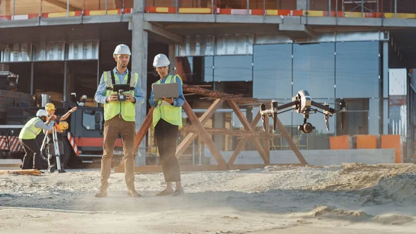 Dos especialistas controlando drones en un sitio de construcción. Ingeniero de arquitectura e inspector de ingeniería de seguridad vuelan dron en construcción de edificios comerciales sitio de control de diseño y calidad — Foto de Stock