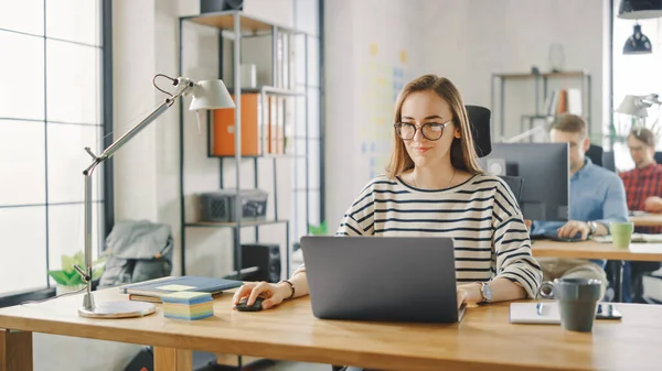 Gyönyörű fiatal nő szemüvegben egy laptopon dolgozik egy kreatív üzleti ügynökségnél. A Loft irodában dolgoznak. Sokszínű emberek dolgoznak a háttérben. Barátságos hangulatban van.. — Stock Fotó