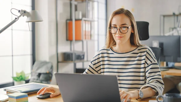 Schöne junge Frau mit Brille arbeitet in einer Kreativagentur an einem Laptop. Sie arbeiten im Loft Office. Diverse Menschen, die im Hintergrund arbeiten. Geschäfte gut gelaunt. — Stockfoto