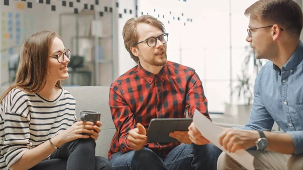 Mujer joven y dos colegas varones teniendo una reunión amistosa y discutiendo nuevas ideas de negocios. Ambiente de Coworking Easygoing en Loft Office Creative Agency. Miran Papeles y Tablet. — Foto de Stock