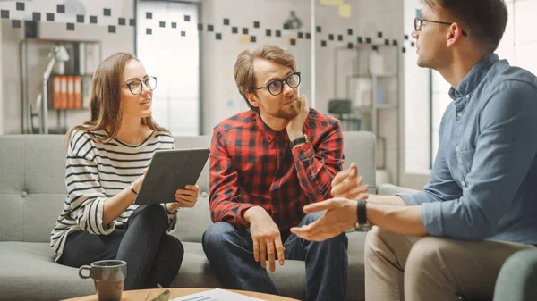 Mujer joven y dos colegas varones teniendo una reunión amistosa y discutiendo nuevas ideas de negocios. Ambiente de Coworking Easygoing en Loft Office Creative Agency. Miran Papeles y Tablet. — Foto de Stock