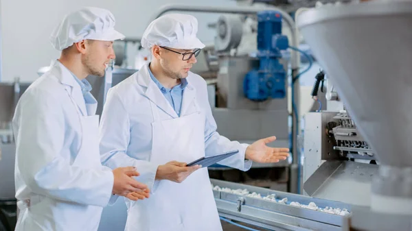 Dois jovens supervisores da qualidade masculina ou técnicos de alimentos estão inspecionando a produção automatizada em uma fábrica de alimentos de despejo. Empregado usa um computador tablet para o trabalho. Eles usam barras brancas de trabalho . — Fotografia de Stock