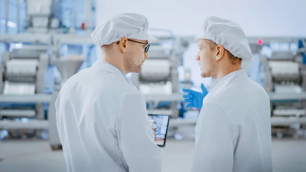 Dos jóvenes empleados de la fábrica de alimentos discuten asuntos relacionados con el trabajo. El Técnico Masculino o Gerente de Calidad utiliza una Tablet Computer para el Trabajo. Llevan sombrero sanitario blanco y ropas de trabajo. —  Fotos de Stock