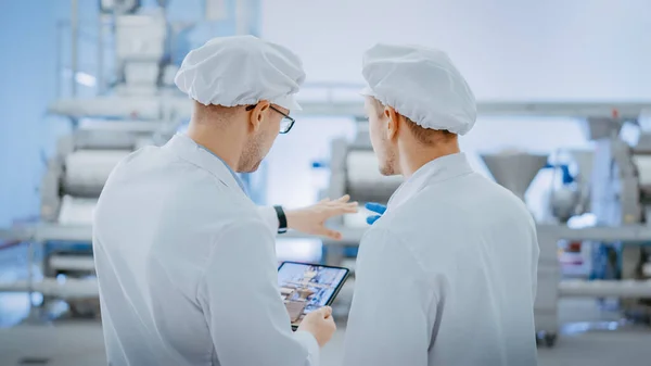 Twee jonge medewerkers van de Food Factory discussiëren over werkgerelateerde zaken. Man Technicus of Quality Manager maakt gebruik van een tablet computer voor het werk. Ze dragen witte badmuts en werkkleding.. — Stockfoto