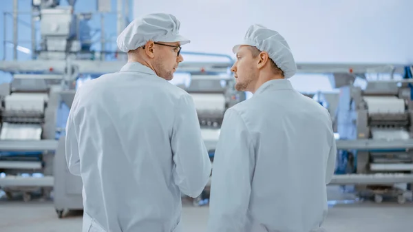 两名年轻食物制造厂雇员讨论与工作有关的事宜。男技师或质量经理使用平板电脑工作。他们穿着白色卫生帽和工作服. — 图库照片