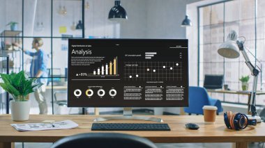 Yaratıcı Modern Ofiste Bir Masaüstü Bilgisayarının Çekimi. Monitör Ekranı Şirket Gelişim Verilerini Grafikler, Grafikler, Yazılım UI ile gösterir. Arka planda Genç Profesyonel Bir Kitap Arıyor.