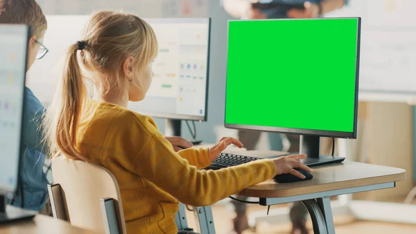 Elementary School Computer Science Classroom: Cute Little Girl Uses Green Mock-up Screen Computer while Learning Coding and Programming. Schoolchildren Getting Modern Education. Over the Shoulder — Stock Photo, Image