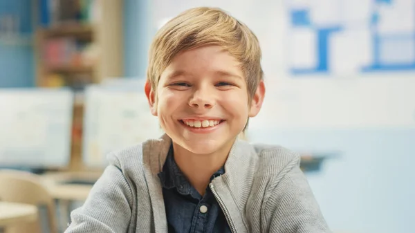 Retrato de un lindo niño sentado en su escritorio de la escuela, ríe felizmente. Niño inteligente con una sonrisa encantadora sentado en el aula. —  Fotos de Stock