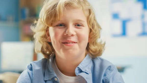 Okul masasında oturan kıvırcık sarı saçlı şirin bir çocuğun portresi mutlu bir şekilde gülümsüyor. Yakışıklı Gülümseyen Akıllı Küçük Çocuk Sınıfta Oturuyor. Yakın çekim. — Stok fotoğraf