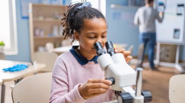 Portrait of Smart Little Schoolgirl Looking Under the Microscope. In Elementary School Classroom Cute Girl Uses Microscope. STEM science, technology, engineering and mathematics Education Program clipart