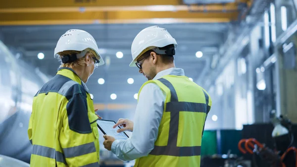 Deux ingénieurs de l'industrie lourde se tiennent dans l'usine de fabrication de tuyaux, utilisent un ordinateur tablette numérique, discutent. Grand tuyau assemblé. Conception et construction de pipelines de transport de pétrole, de gaz et de combustibles — Photo