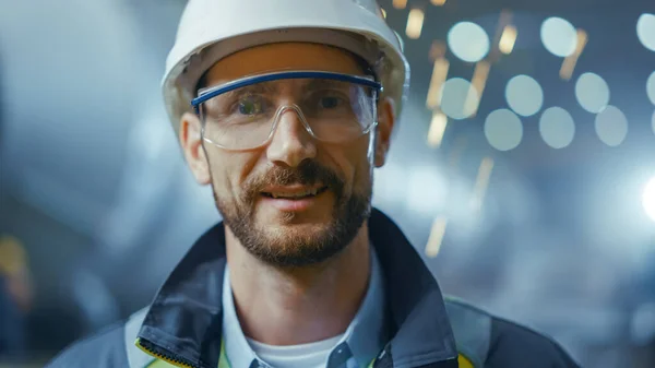 Portrait d'un ingénieur professionnel de l'industrie lourde portant uniforme de sécurité, lunettes et casque rigide souriant. En arrière-plan Grande usine industrielle non ciblée où les étincelles de soudage volent — Photo