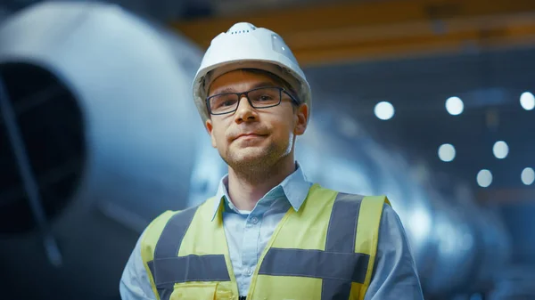 Porträtt av ung professionell tung industri ingenjör arbetare bär säkerhetsväst och hårdhatt leende på kameran. I bakgrunden ofokuserad stor industrifabrik där svetsgnistor flyger. — Stockfoto