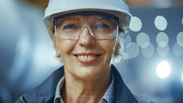 Retrato de una ingeniera profesional de la industria pesada que usa uniforme de seguridad y sombrero duro, gafas y sonrisas encantadoramente. En el fondo Fábrica industrial desenfocada donde la soldadura chispa volando —  Fotos de Stock