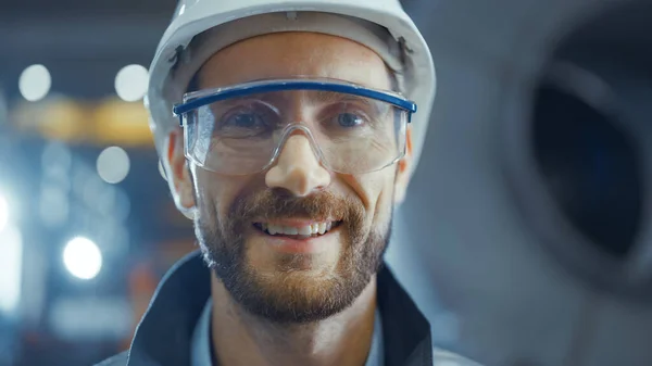 Porträt eines lächelnden, professionellen Schwerindustrie-Ingenieurs, der Sicherheitsuniform, Brille und Hut trägt. Im Hintergrund unkonzentrierte große Industriefabrik — Stockfoto