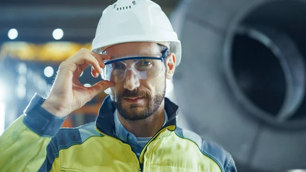 Porträt eines lächelnden, professionellen Schwerindustrie-Ingenieurs, der Sicherheitsuniform trägt und eine Brille aufsetzt. Im Hintergrund unkonzentrierte große Industriefabrik — Stockfoto