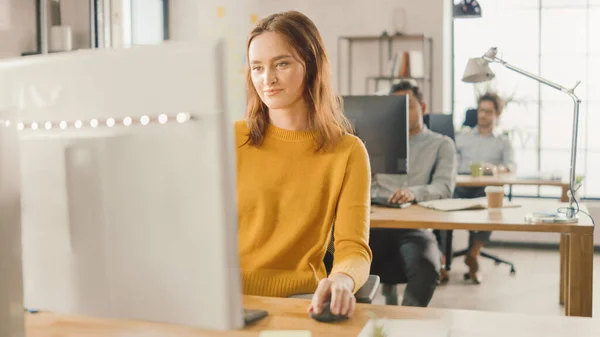 Bella e intelligente dai capelli rossi specialista femminile seduta alla sua scrivania lavora su un computer desktop. Sullo sfondo il moderno ufficio luminoso con diversi gruppi di professionisti che lavorano per la crescita delle startup — Foto Stock