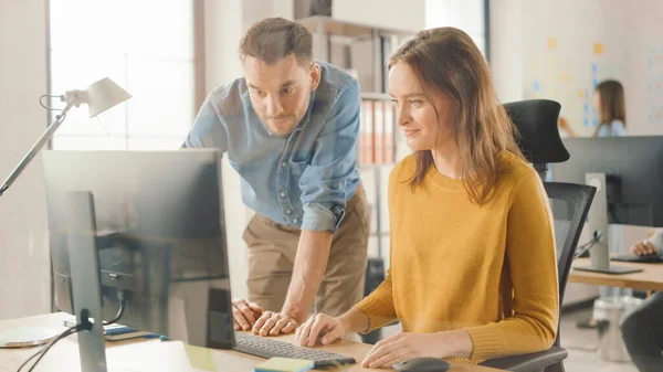 Especialista feminina trabalha no computador desktop, gerente de projeto está ao lado e dá conselhos sobre a otimização do fluxo de trabalho para a gestão da experiência do cliente. Escritório moderno com equipe diversa de profissionais — Fotografia de Stock