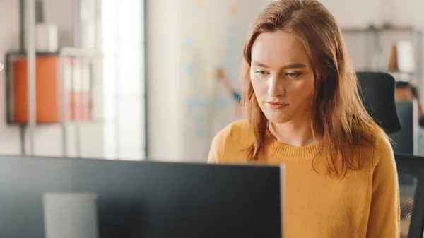 Die schöne und kluge rothaarige Spezialistin sitzt an ihrem Schreibtisch und arbeitet an einem Desktop-Computer. Helles und modernes Großraumbüro mit stylischen, ergonomischen Möbeln für talentierte Kreative — Stockfoto