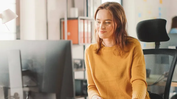 Die schöne und kluge rothaarige Spezialistin sitzt an ihrem Schreibtisch und arbeitet an einem Desktop-Computer. Helles und modernes Großraumbüro mit stylischen, ergonomischen Möbeln für talentierte Kreative — Stockfoto