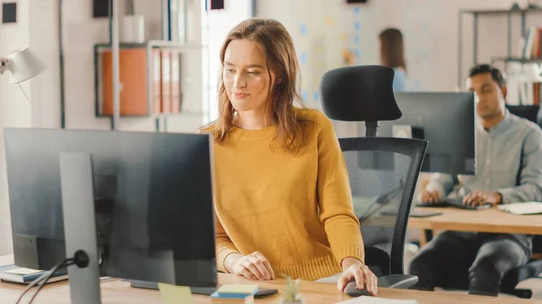 Gyönyörű és okos vörös hajú nő specialista ül az asztalánál dolgozik egy asztali számítógép. Fényes és modern Open Space Office stílusos ergonomikus bútorok a tehetséges kreatív emberek — Stock Fotó