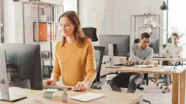 Linda e inteligente Red Haired Especialista feminina sentada em sua mesa funciona em um computador desktop. No fundo Escritório Brilhante Moderno com Grupo Diverso de Profissionais Trabalhando para Crescer Startup — Fotografia de Stock