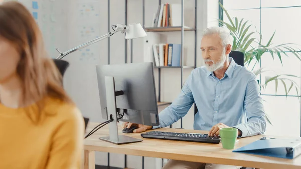 Handsome Senior Office Employee Sitting at His Desk Works on a Desktop Computer. In the Background Modern Bright Office with Diverse Group of Professionals Working for Growing Startup