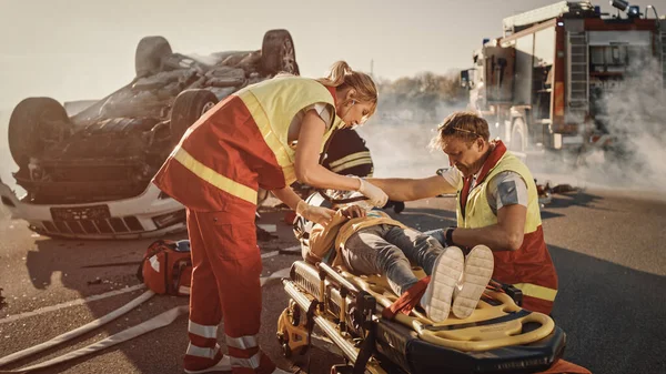 En la escena del accidente de tráfico: Paramédicos salvando la vida de una niña mintiendo en camillas, le han dado primeros auxilios. El bombero lleva a la madre a la hija. En el vehículo de vuelco de fondo — Foto de Stock