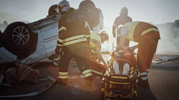 交通事故現場:消防救助隊が女性被害者をロールオーバー車両から引き抜き、ストレッチャーを慎重に使用し、応急処置を行う救急隊員に引き渡す — ストック写真