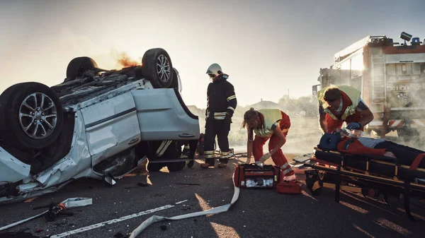 O dopravní nehodě při autonehodě: Záchranáři a hasiči Zachraňte zraněné oběti v pasti. Doktoři dávají první pomoc ženám na nosítkách. Hasiči použít hydraulické řezačky rozmetadlo k otevření vozidla — Stock fotografie