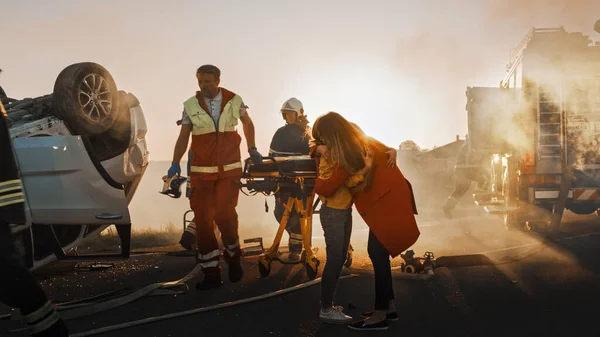 Madre ferita e giovane figlia si riuniscono dopo terribile incidente stradale d'auto, si abbracciano felicemente. Sullo sfondo attraverso fumo e fuoco, coraggiosi paramedici e vigili del fuoco salvano vite umane — Foto Stock