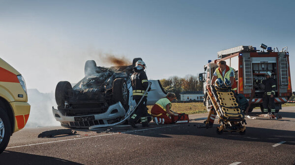 On the Car Crash Traffic Accident Scene: Team of Firefighters Rescue Injured People Trapped in Rollover Vehicle. Professionals Extricate Victims, give First Aid, Extinguish Fire